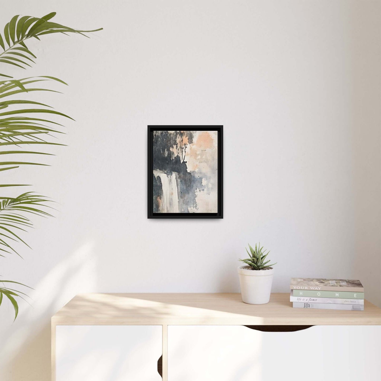 Framed waterfall art print on matte canvas decorating a minimalist room with a plant, books, and light wooden surface.