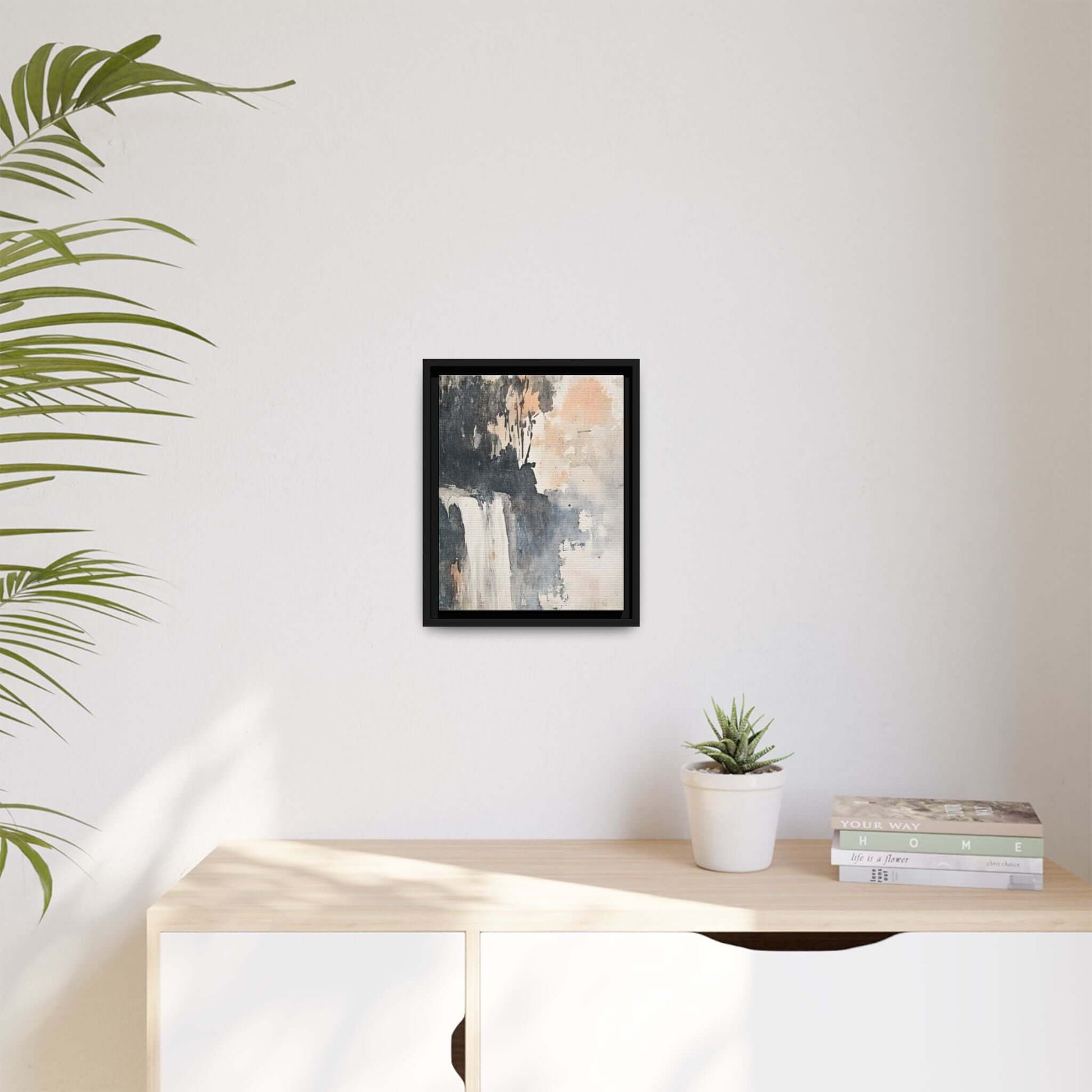 Framed waterfall art print on matte canvas decorating a minimalist room with a plant, books, and light wooden surface.