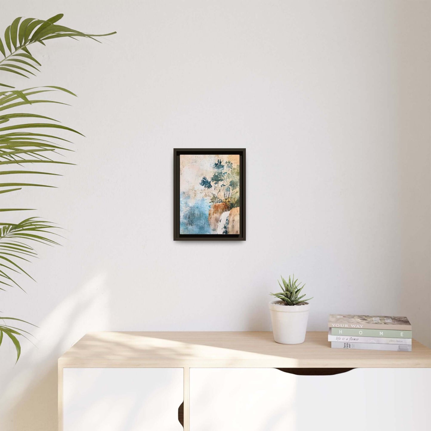Framed watercolor canvas print of a misty forest landscape on a white wall above a light wooden cabinet with books and a plant.