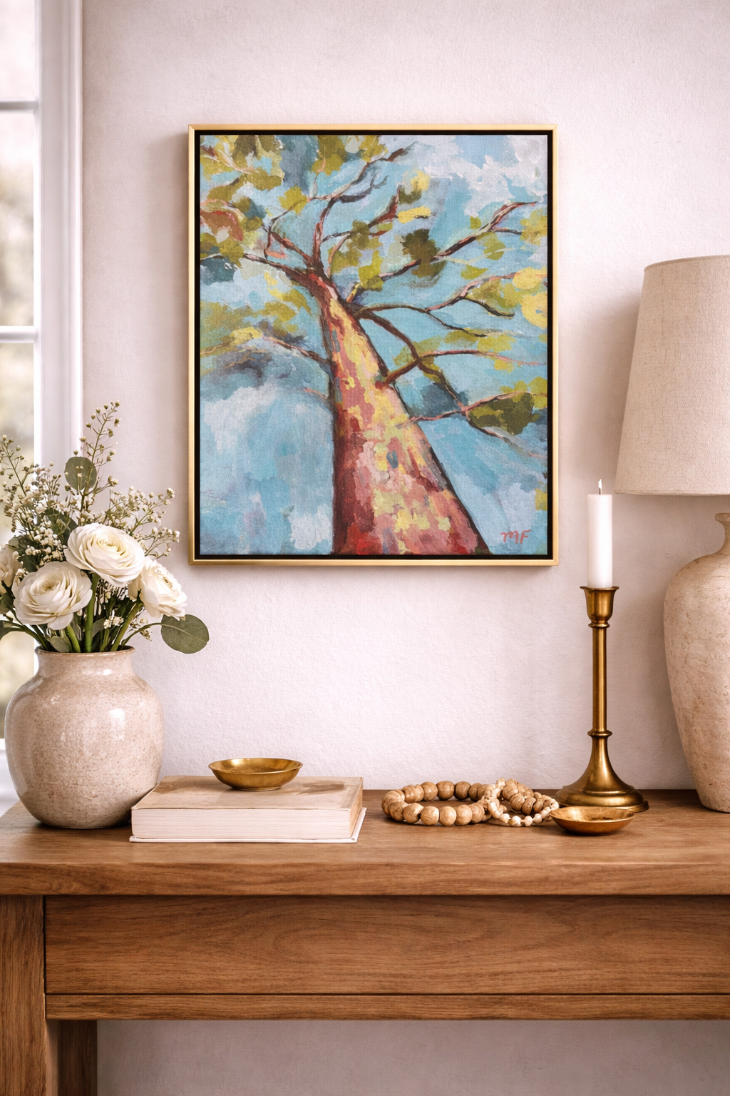 Decorative setup with a painting of a tree, vase with flowers, books, and lamp on a wooden surface.