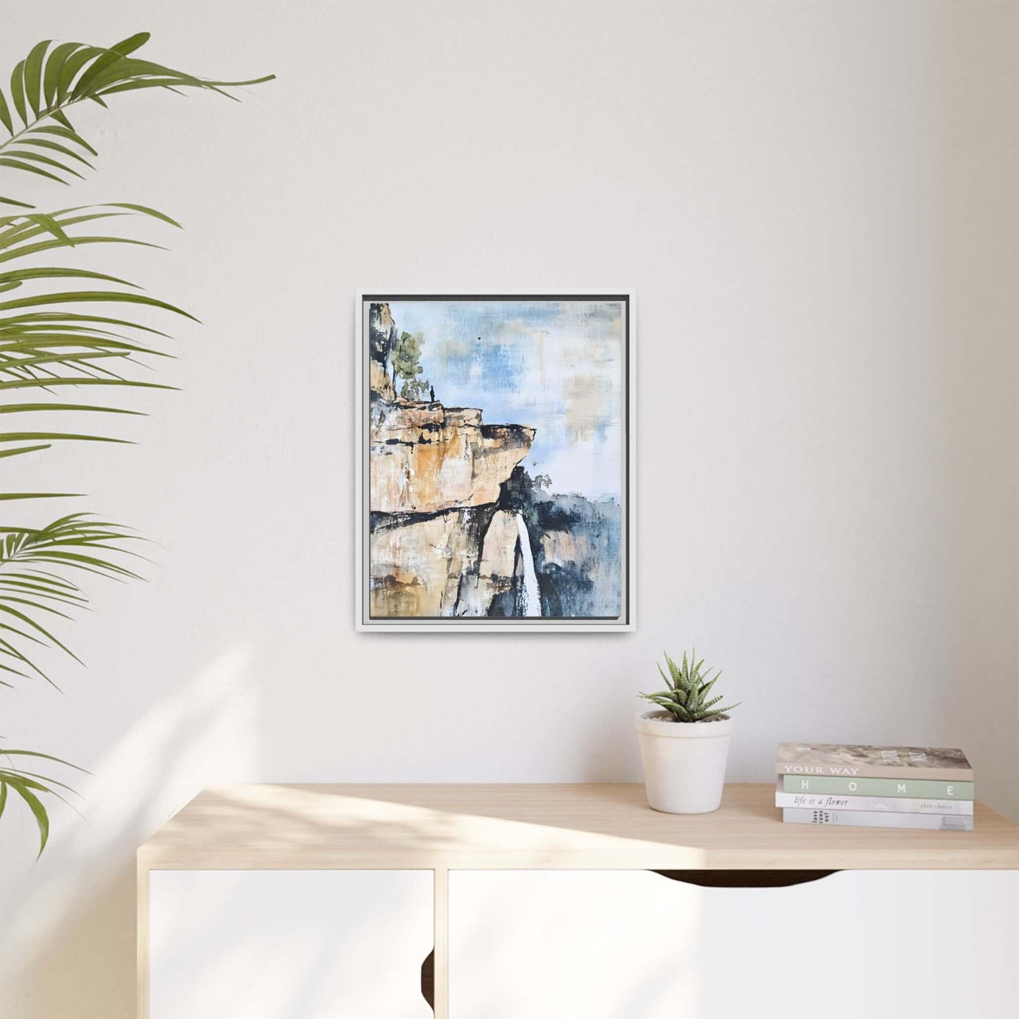Framed impressionistic scenic landscape waterfall canvas wall art hung on a minimalist room wall beside a plant and books.