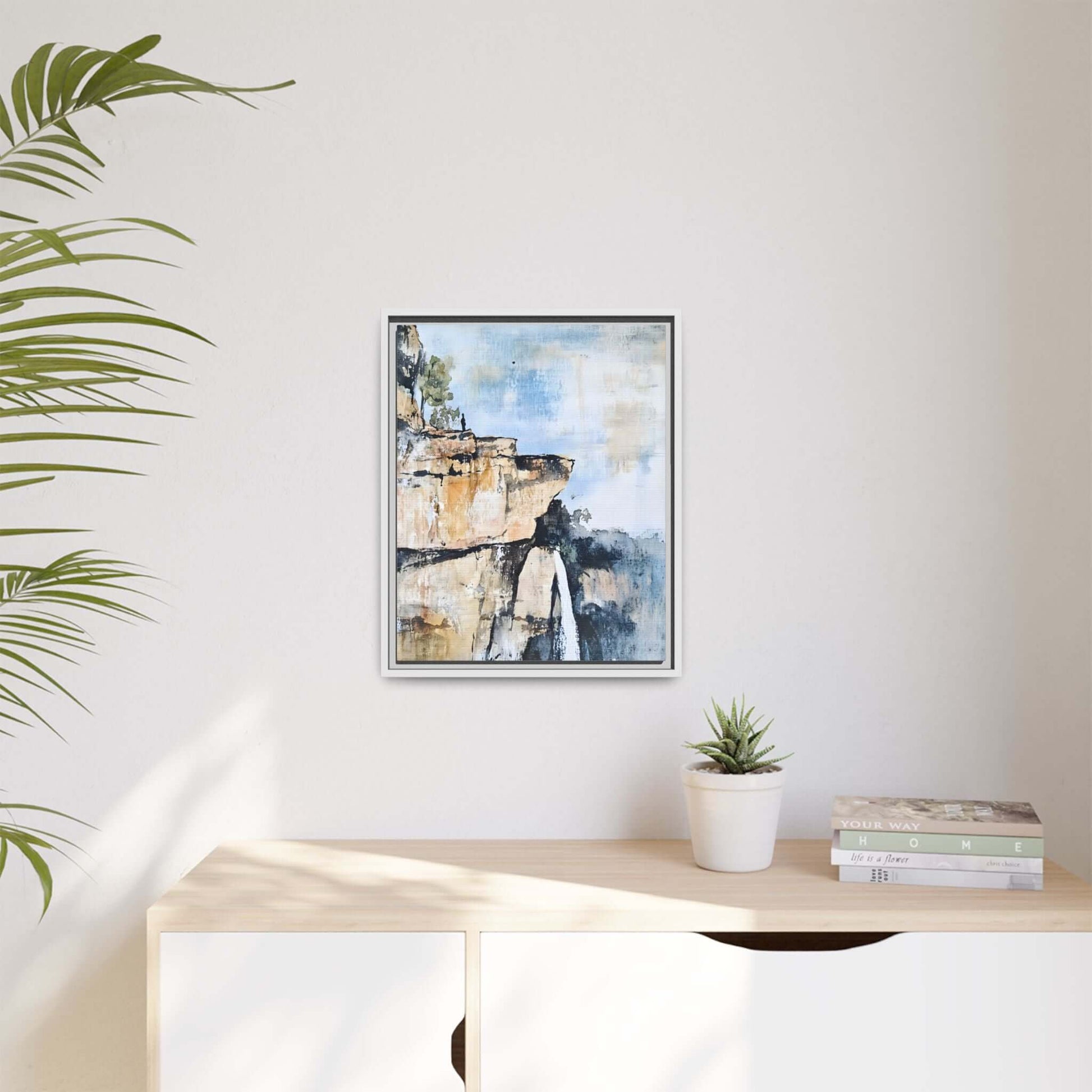 Framed impressionistic scenic landscape waterfall canvas wall art hung on a minimalist room wall beside a plant and books.