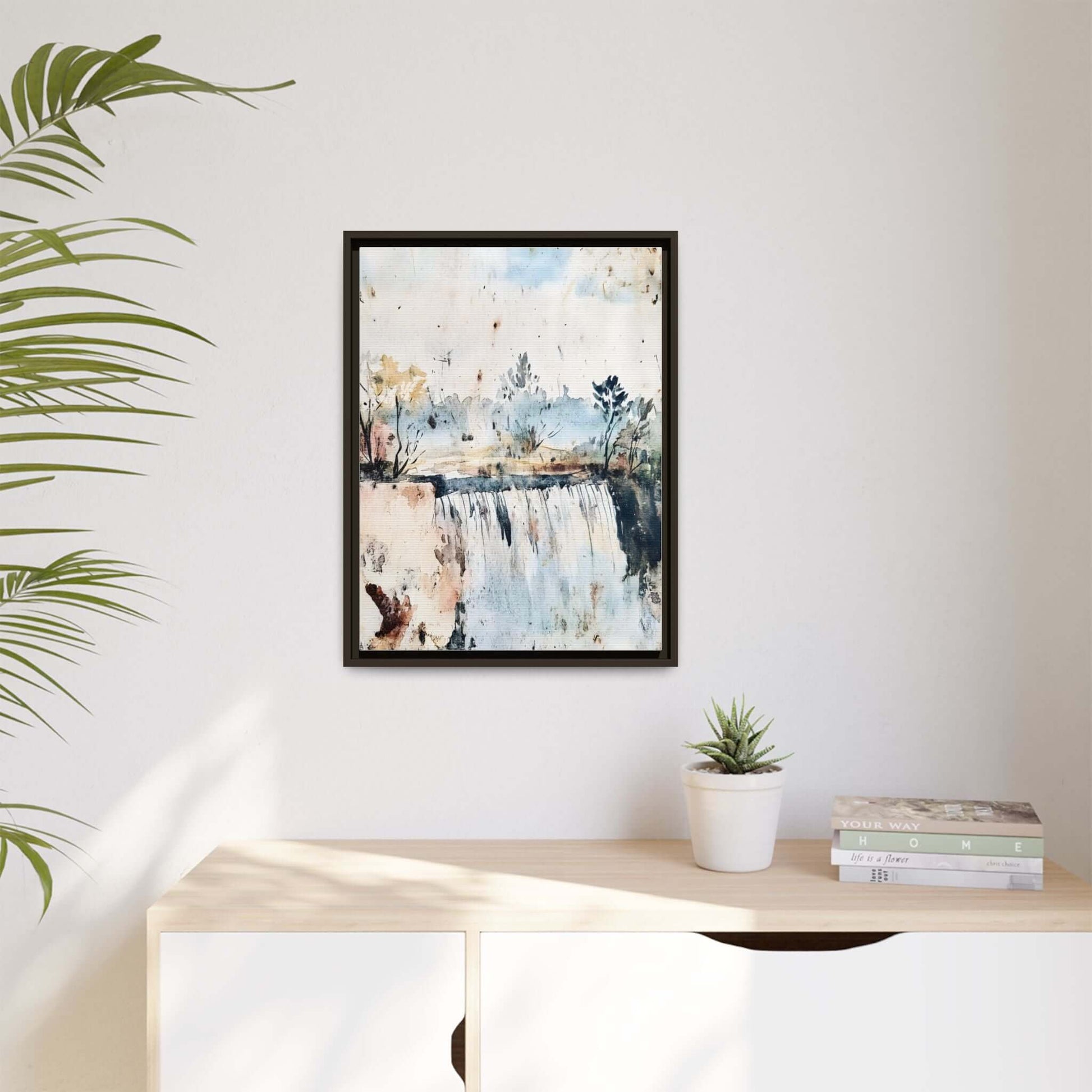 Framed watercolor wall art depicting a serene landscape with a waterfall, displayed in a modern room with a potted plant and books.