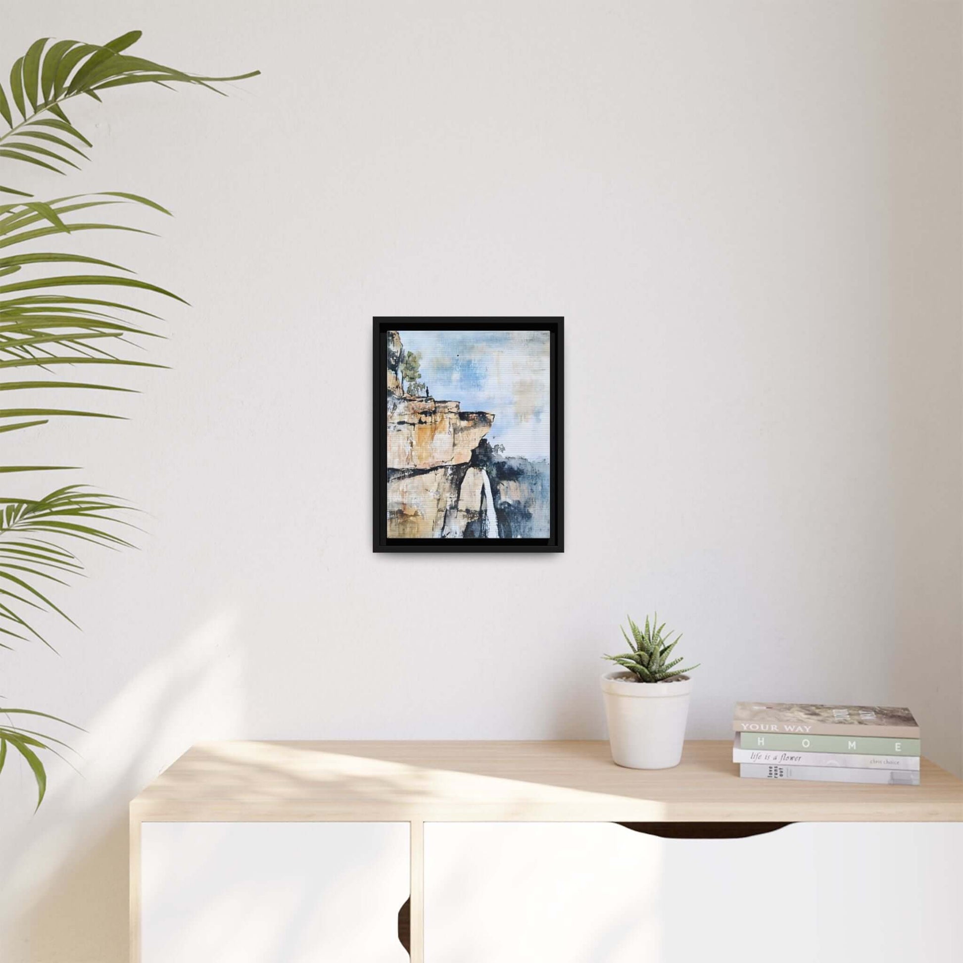Framed impressionistic scenic landscape waterfall canvas wall art displayed above light wood cabinet with plant and books.