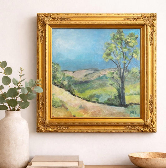 Framed painting of a landscape with a tree on a wall next to a vase and books.